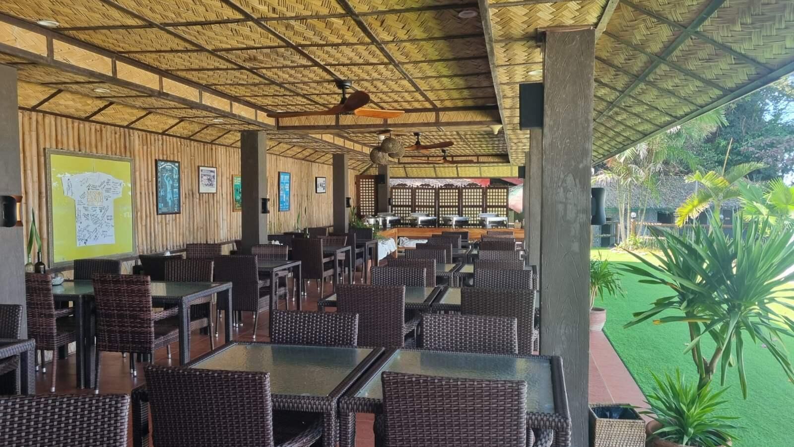 Un restaurante al aire libre con mesas y sillas de mimbre, techo de bambú y un césped verde visible a través de sus espacios abiertos.