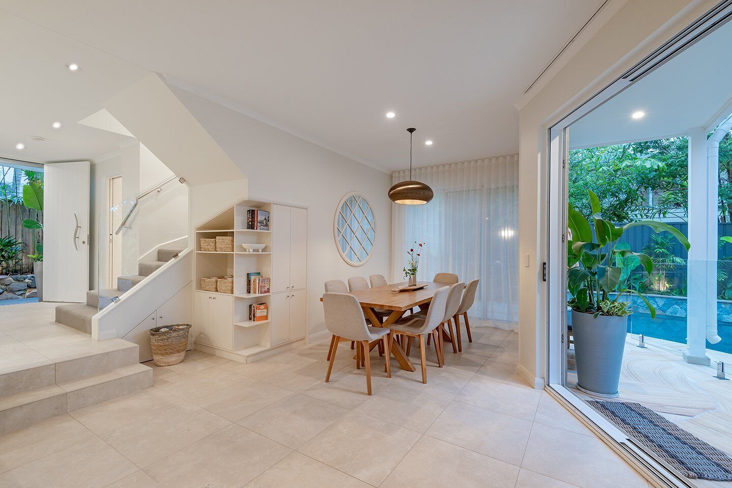 A modern interior with a staircase, a dining area with a wooden table and chairs, and a large window providing a view of greenery outside.
