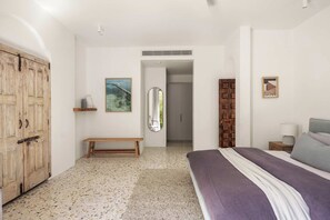 A bedroom with a bed, a wooden wardrobe, a bench, a mirror, and a framed picture on the wall.