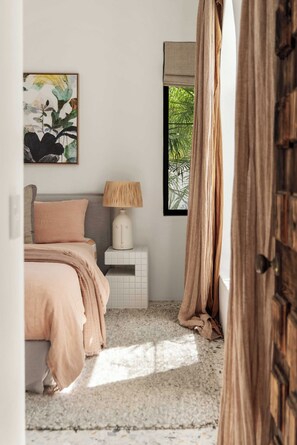 A bedroom with a bed, a nightstand, a lamp, a window with curtains, and a painting on the wall.
