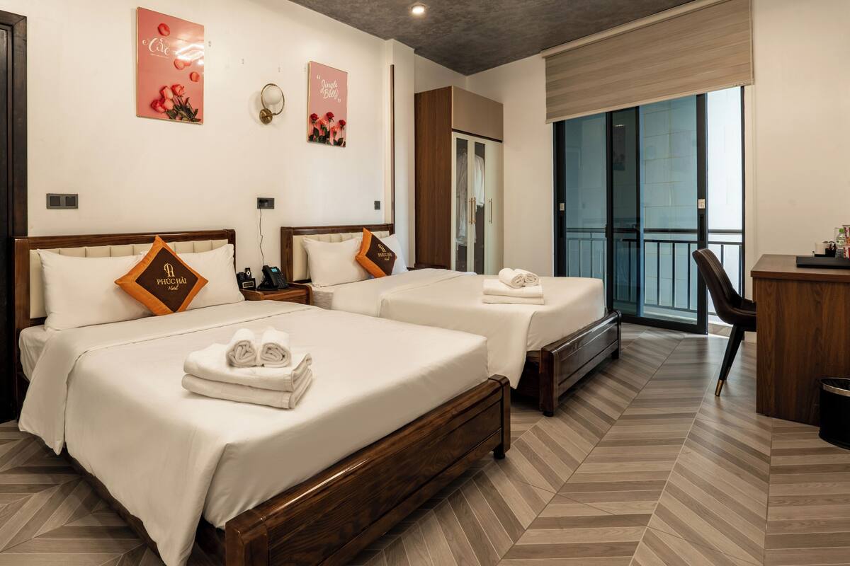 Quarto de hotel com duas camas, uma escrivaninha, uma cadeira e varanda com vista.