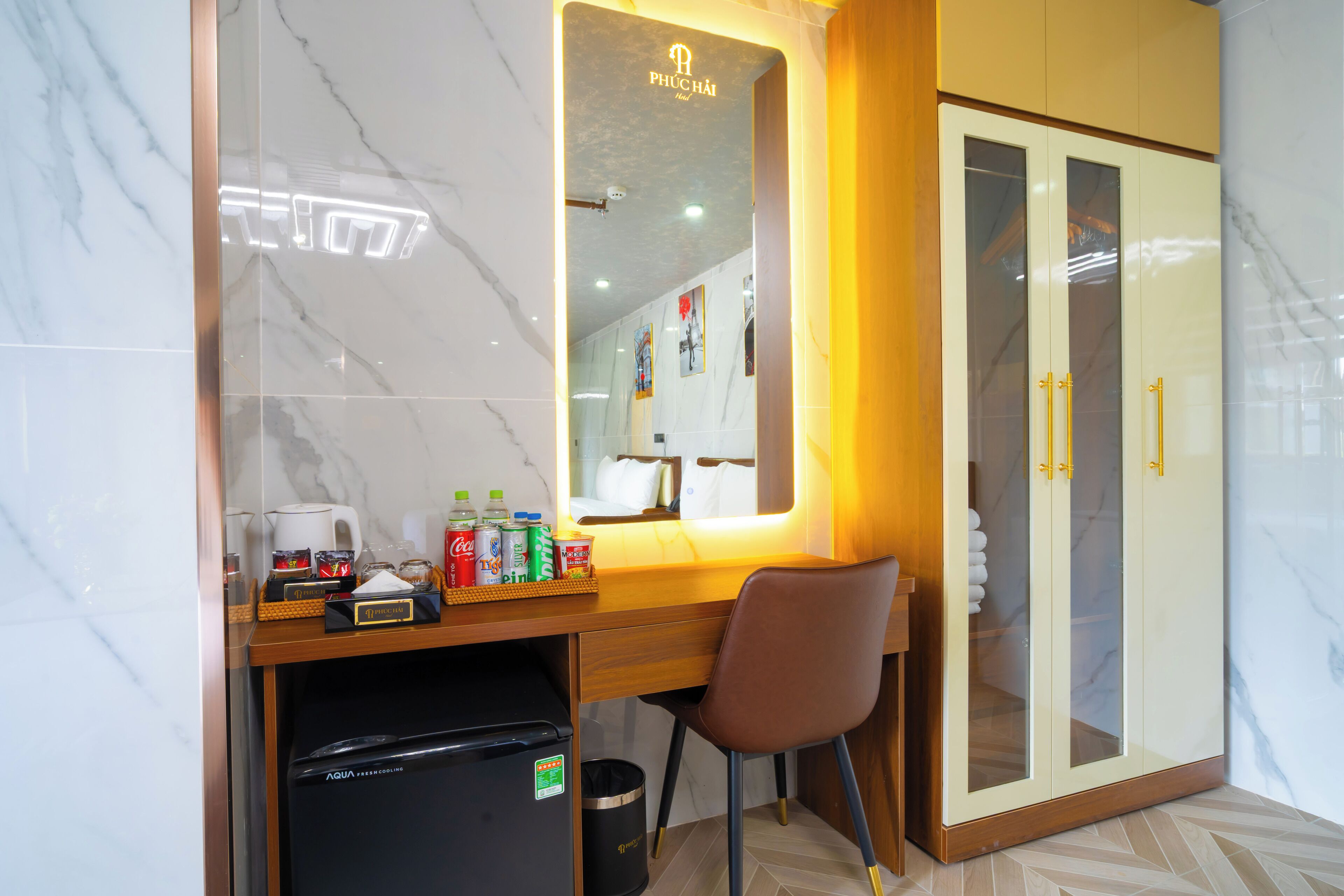 A hotel room with a marble-tiled wall, a wooden desk with a mirror, a brown chair, a wardrobe with glass doors, and a minibar.