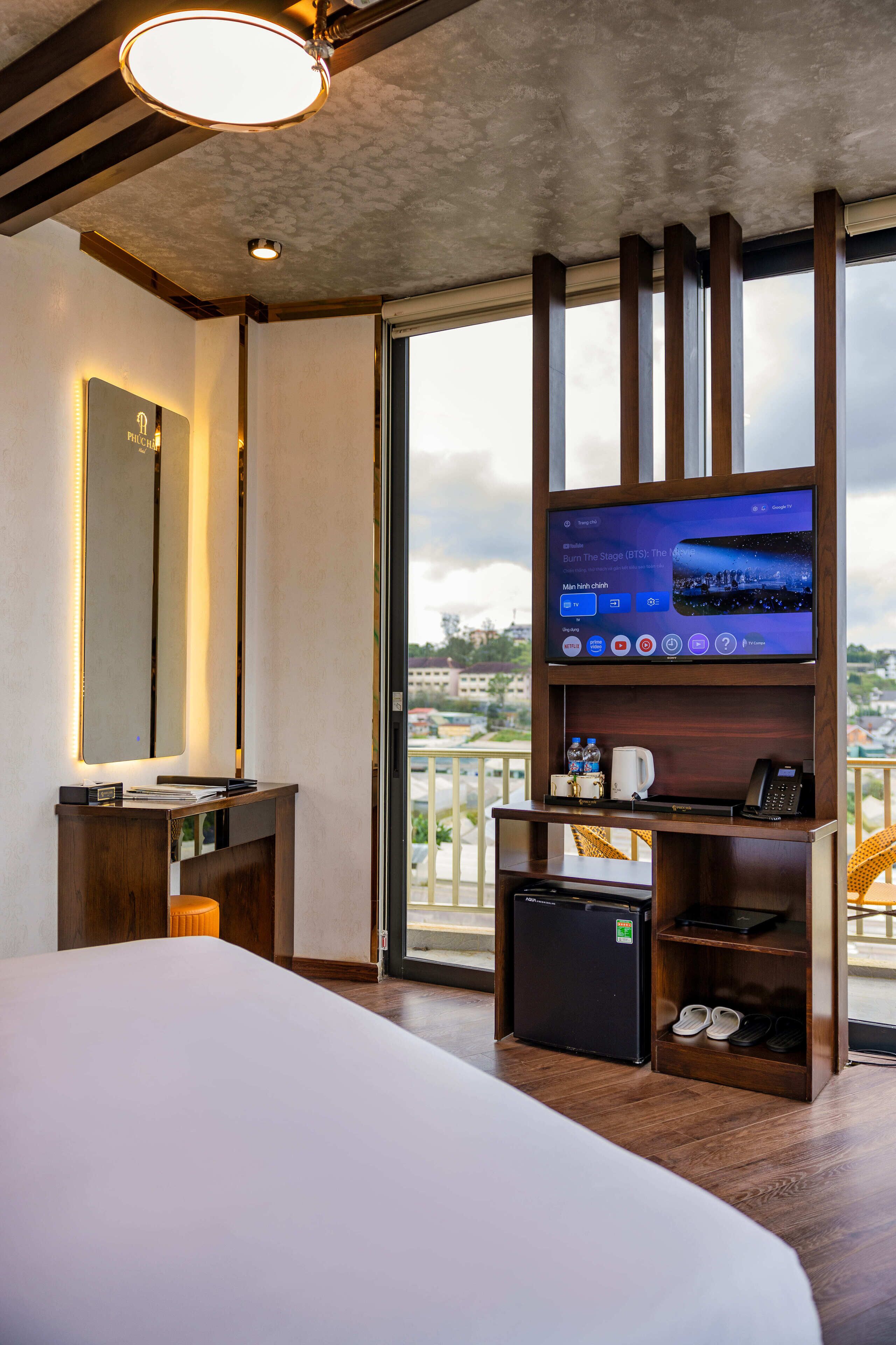 Habitación de hotel con una cama grande, un televisor sobre un soporte de madera, un refrigerador pequeño y un balcón con vistas.