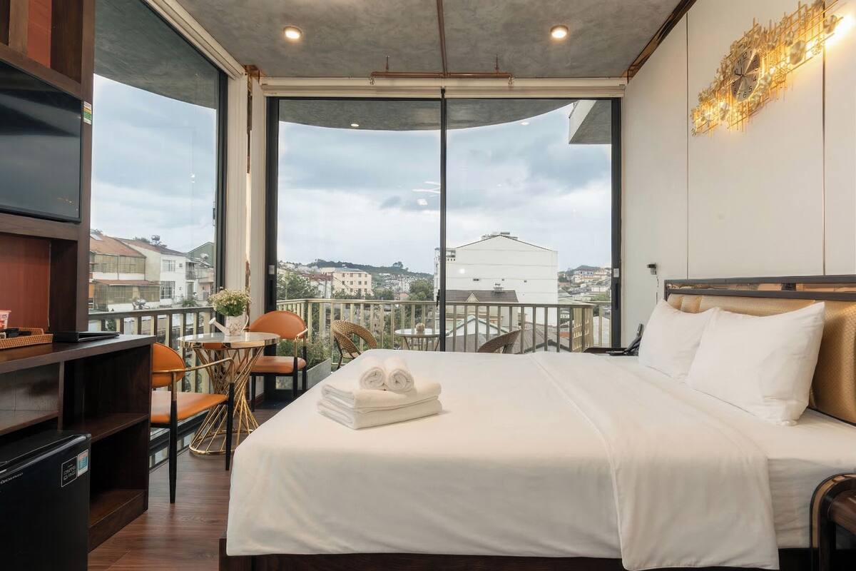 Quarto com cama, televisão, mesa pequena com cadeiras e vista para a cidade.