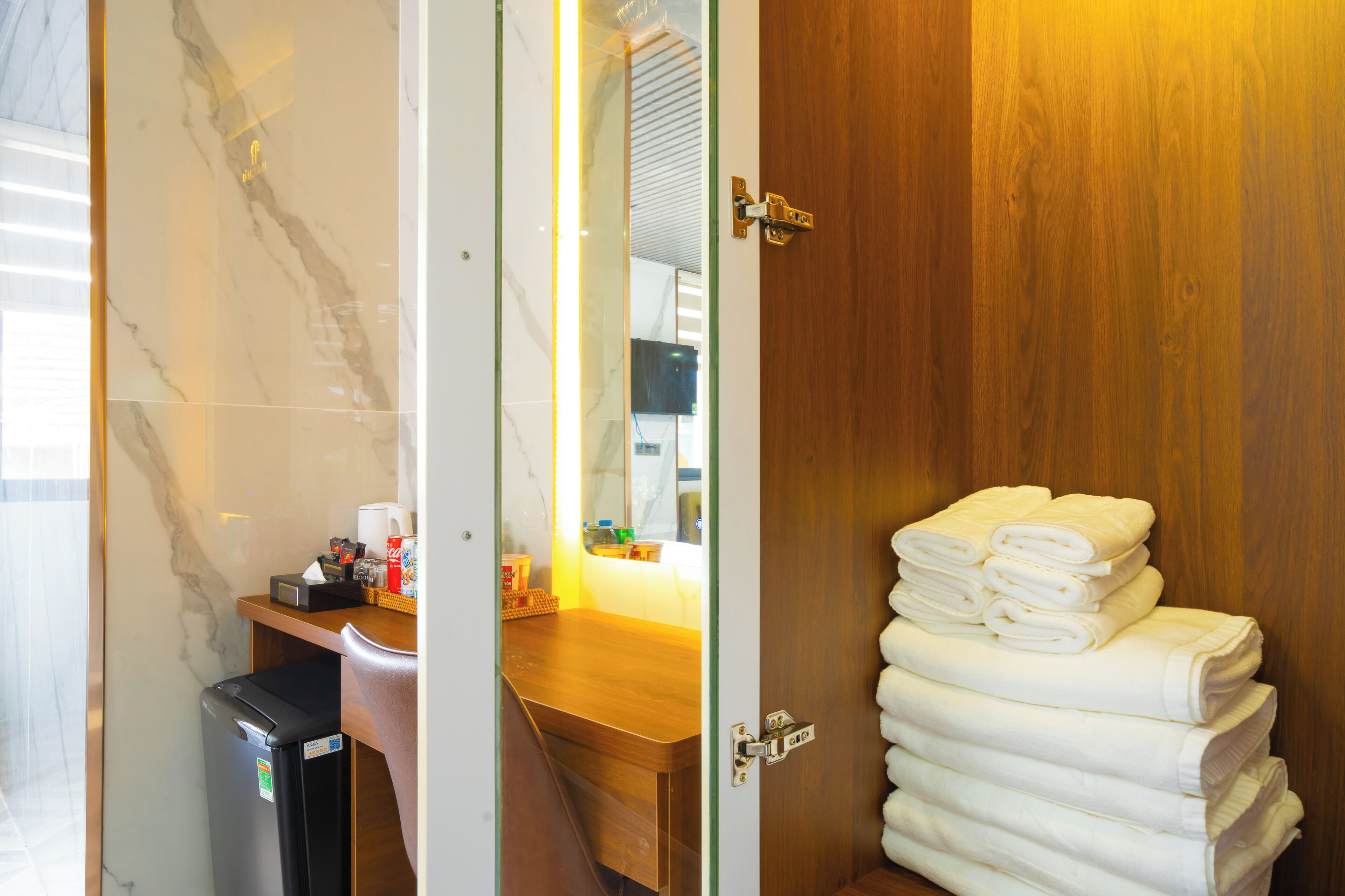 Une chambre d’hôtel avec une porte en bois, un mur en marbre, un comptoir en bois où sont posés des articles de toilette, et une pile de serviettes blanches.