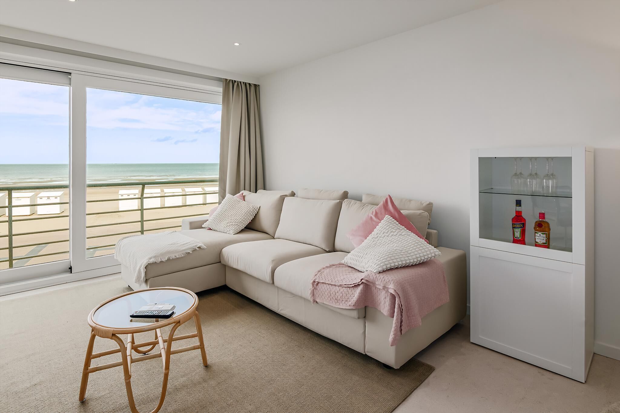 A modern living room with a beige sofa, a glass coffee table, and a view of the beach.