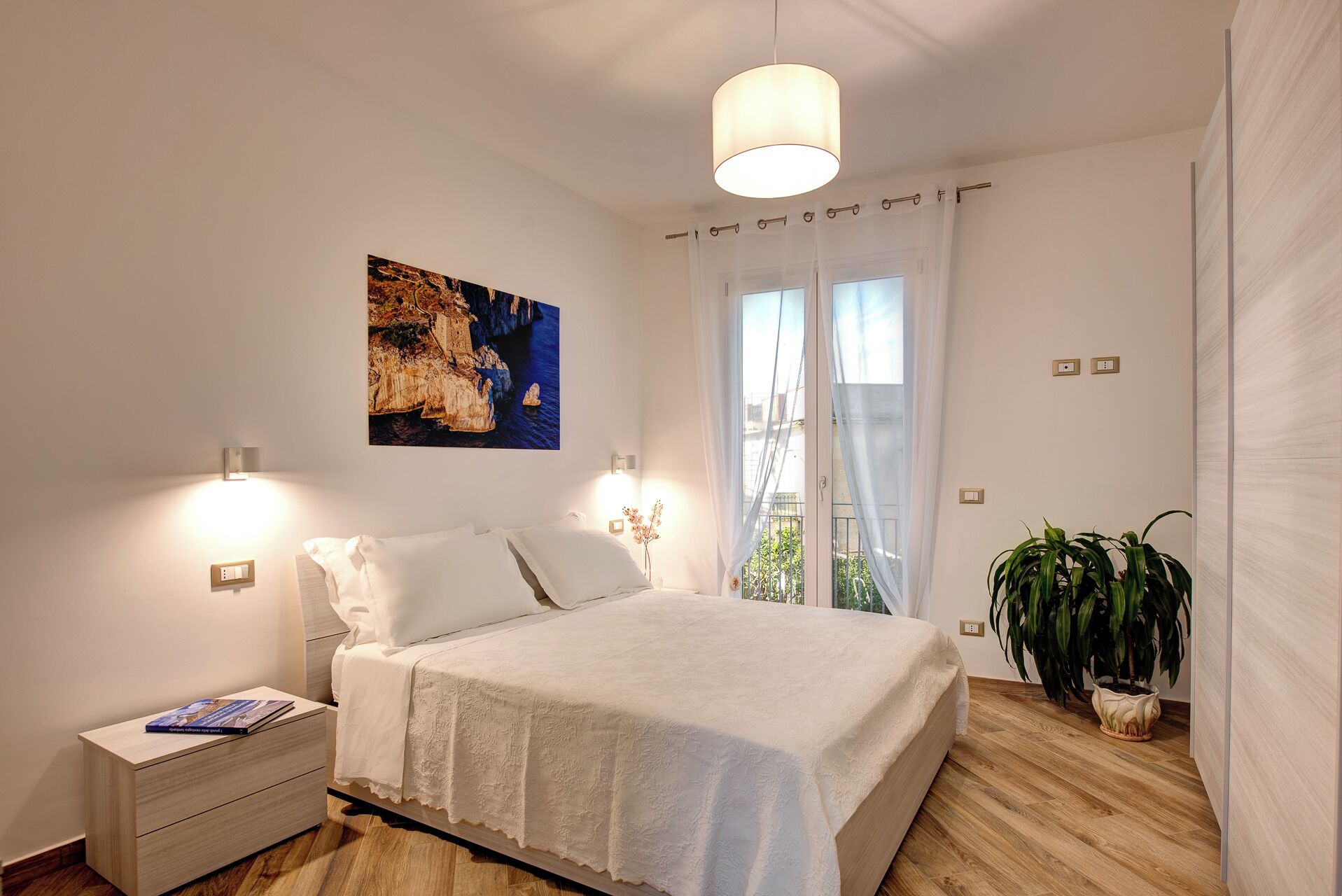 Quarto com cama, mesas de cabeceira, um abajur na parede, janela com cortinas e um vaso com planta.