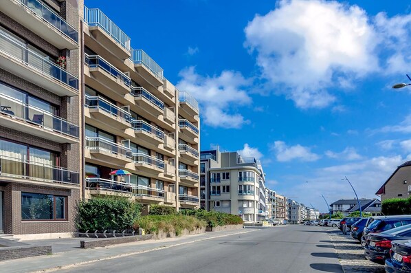 A multi-story residential building with balconies, a parking area, and a clear blue sky.