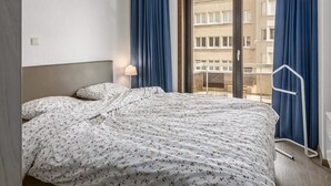 Ein Hotelzimmer mit Bett, Nachttisch, Lampe und Blick auf die Stadt durch ein großes Fenster.