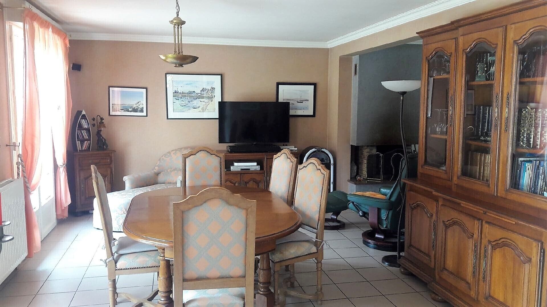 Un comedor con una mesa redonda de madera, sillas tapizadas, un televisor, cuadros enmarcados en la pared y un aparador de madera con puertas de cristal.