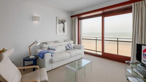 Ein modernes Hotelzimmer mit einem weißen Sofa, einem Glastisch, einem Beistelltisch mit Lampe und einem großen Fenster mit Blick auf den Strand.
