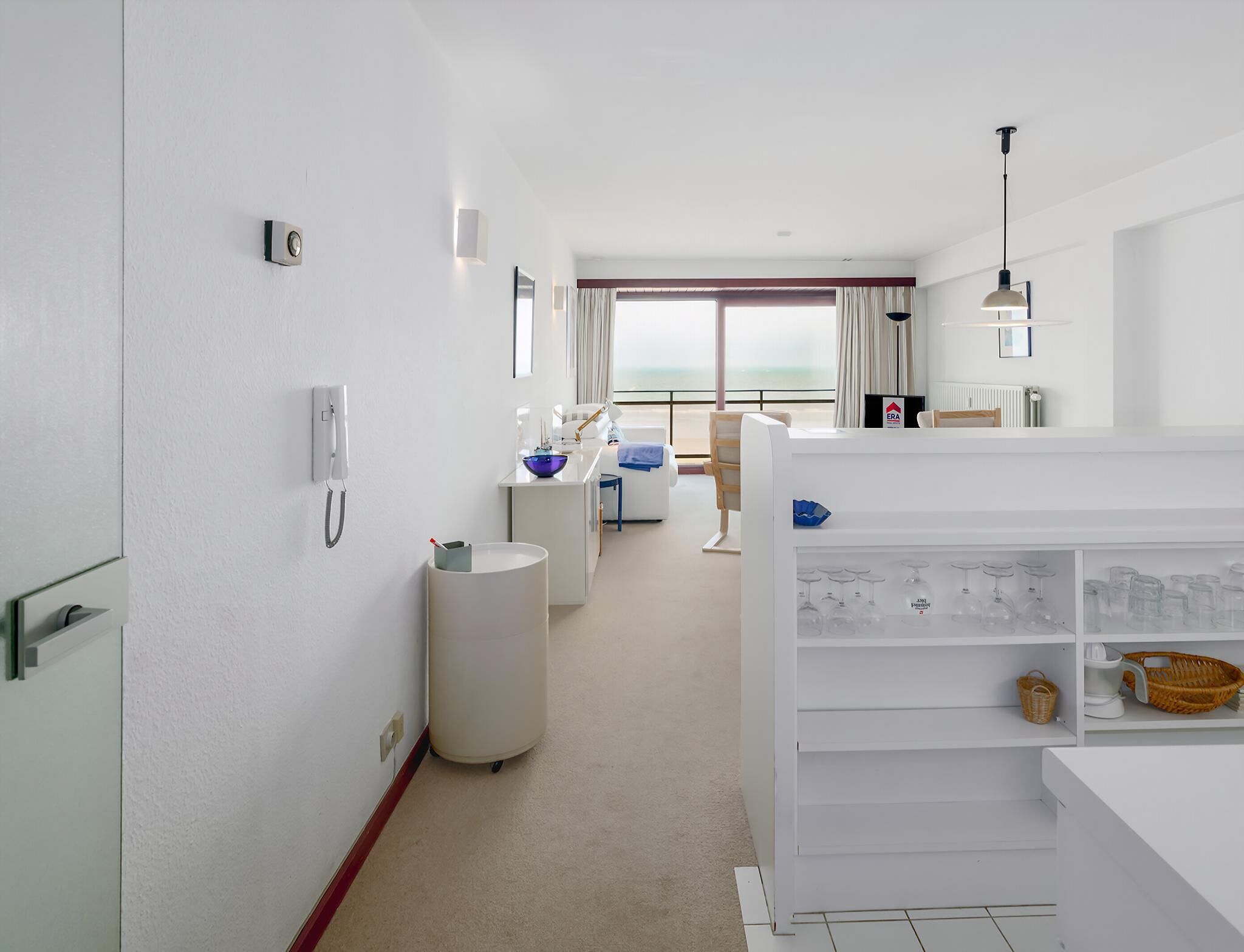 Une chambre d’hôtel moderne dotée d’une grande fenêtre, d’un mini-bar blanc, d’un bureau avec une lampe et offrant une vue sur la mer.