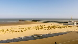 Una playa con un muelle largo que se extiende hacia el mar, una zona arenosa con vegetación escasa y un edificio con una torre.