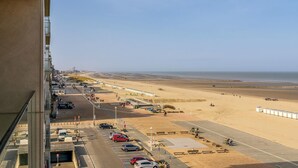 Un estacionamiento frente a la playa con coches, un paseo peatonal y vistas al mar.