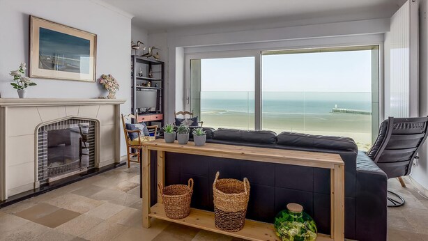 Una sala de estar con chimenea, un ventanal con vista a la playa, un sofá negro y una mesa consola de madera con cestas.