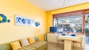 A bright yellow room with a striped sofa, a dining table with chairs, and a large window with a view of trees. - il nido sul Mare by Interhome (Cervo)