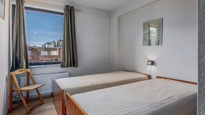 A small, clean room with two single beds, a wooden chair, a window with curtains, a radiator, and a wall-mounted light. - 1 bedroom accommodation in Sint Idesbald (Sint Idesbald)
