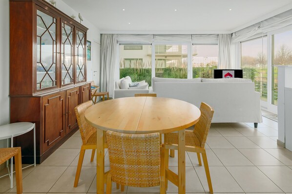 A modern living room with a round wooden dining table, wicker chairs, a white sofa, and a large cabinet with glass doors.