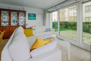 A modern living room with a white sofa, yellow cushions, a dining area with a wooden cabinet, and a large sliding glass door leading to a garden.