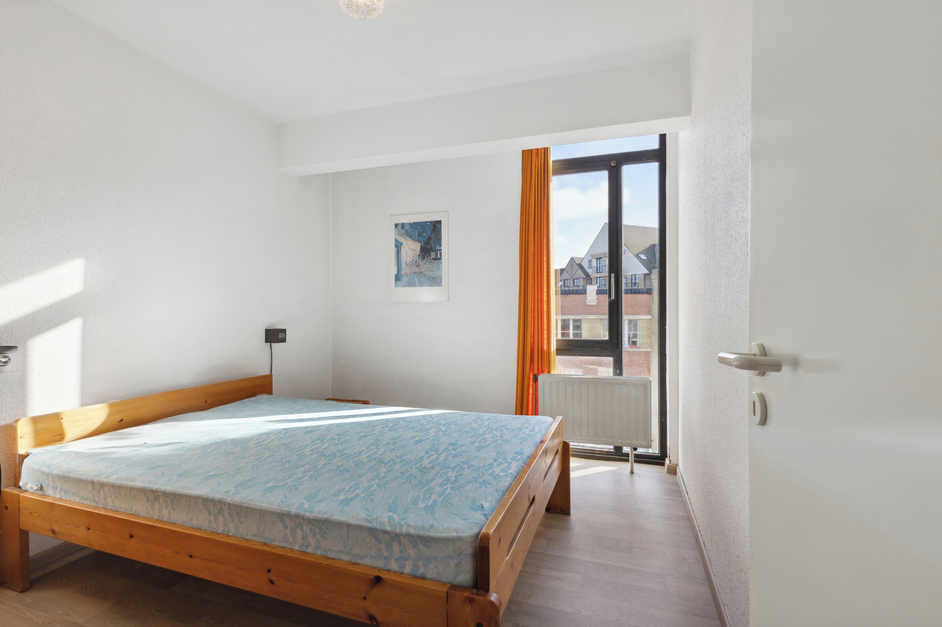 A bedroom with a wooden bed, a radiator, a window with curtains, and a picture on the wall.