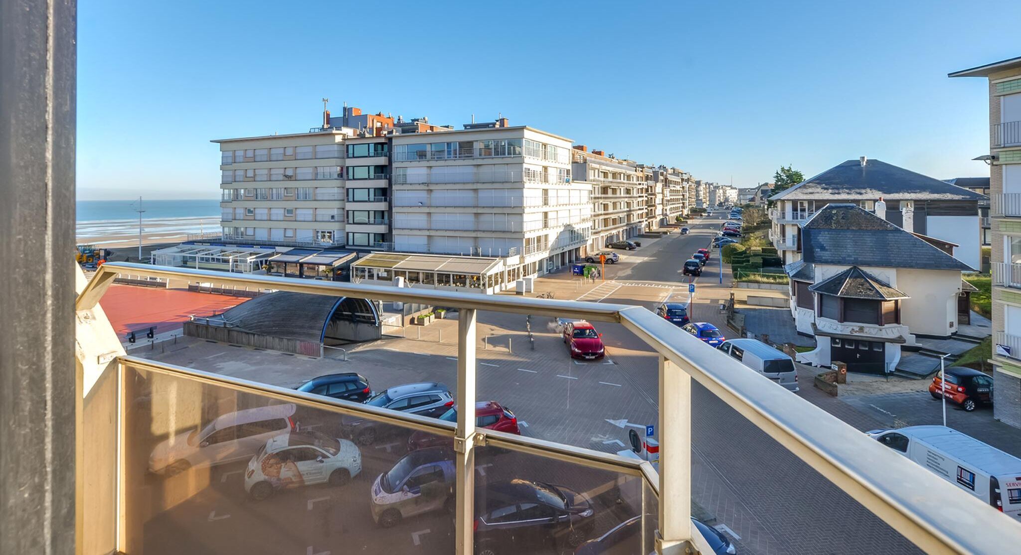 Une vue depuis un balcon donnant sur une rue où sont garées des voitures, avec un passage piéton et des immeubles dotés de balcons.