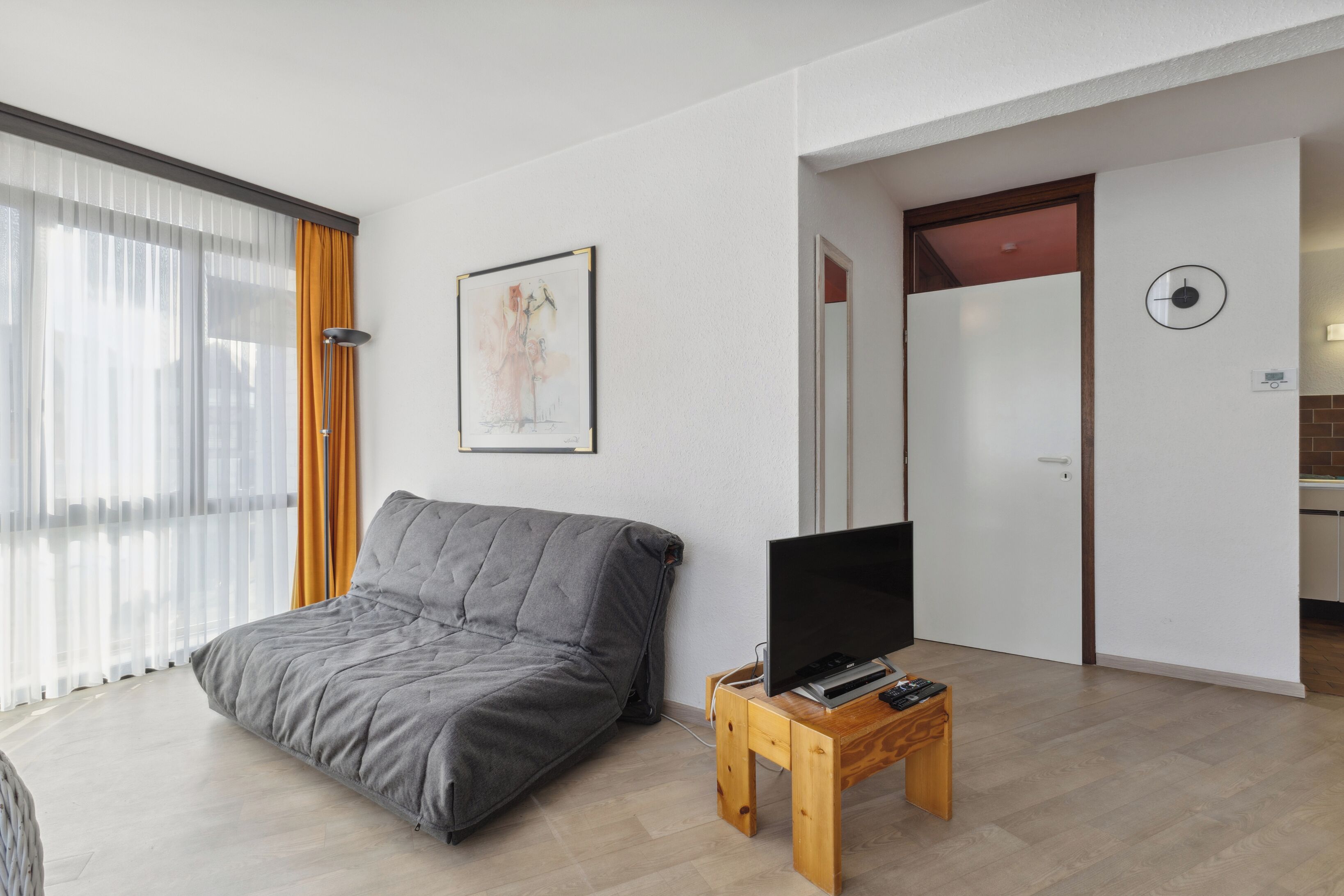 A modern living room with a grey sofa, a flat-screen TV on a wooden stand, a painting on the wall, and a sliding door leading to another room.