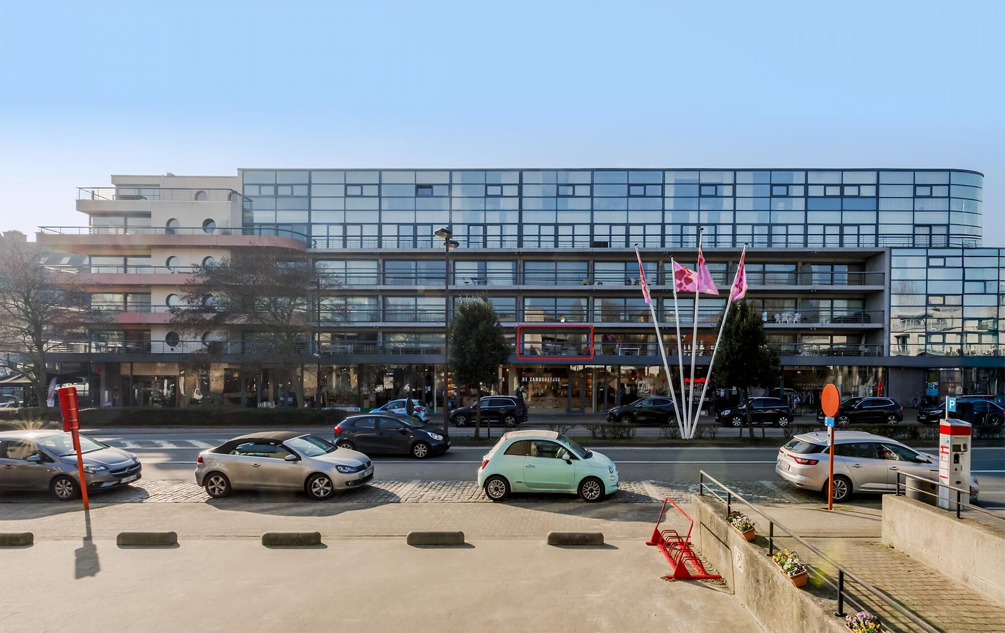Un edificio moderno con fachada de vidrio, varios espacios de estacionamiento y algunos coches aparcados en la entrada.