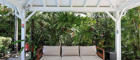 A covered outdoor seating area with a woven hanging light fixture, a cushioned bench with a table, and lush greenery in the background.