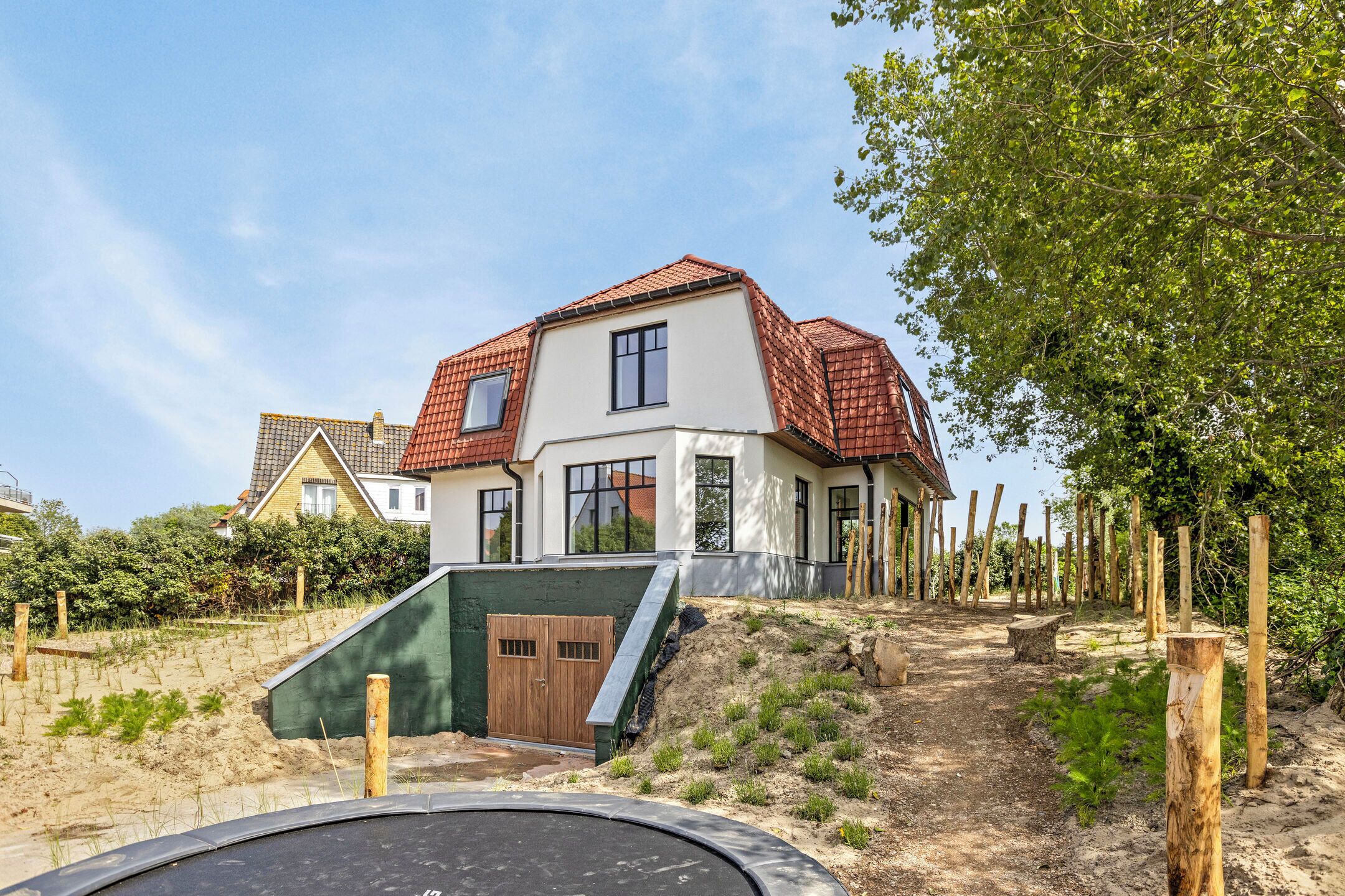 Une maison moderne à deux étages, avec un toit recouvert de tuiles rouges, une porte verte et un petit jardin délimité par une clôture en bois.