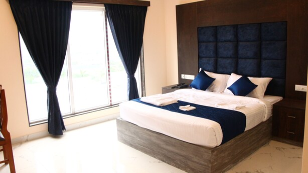 A bedroom with a large bed, a wooden headboard, a nightstand, and a window with curtains. - SAS SQUARE (Tiruchirappalli)