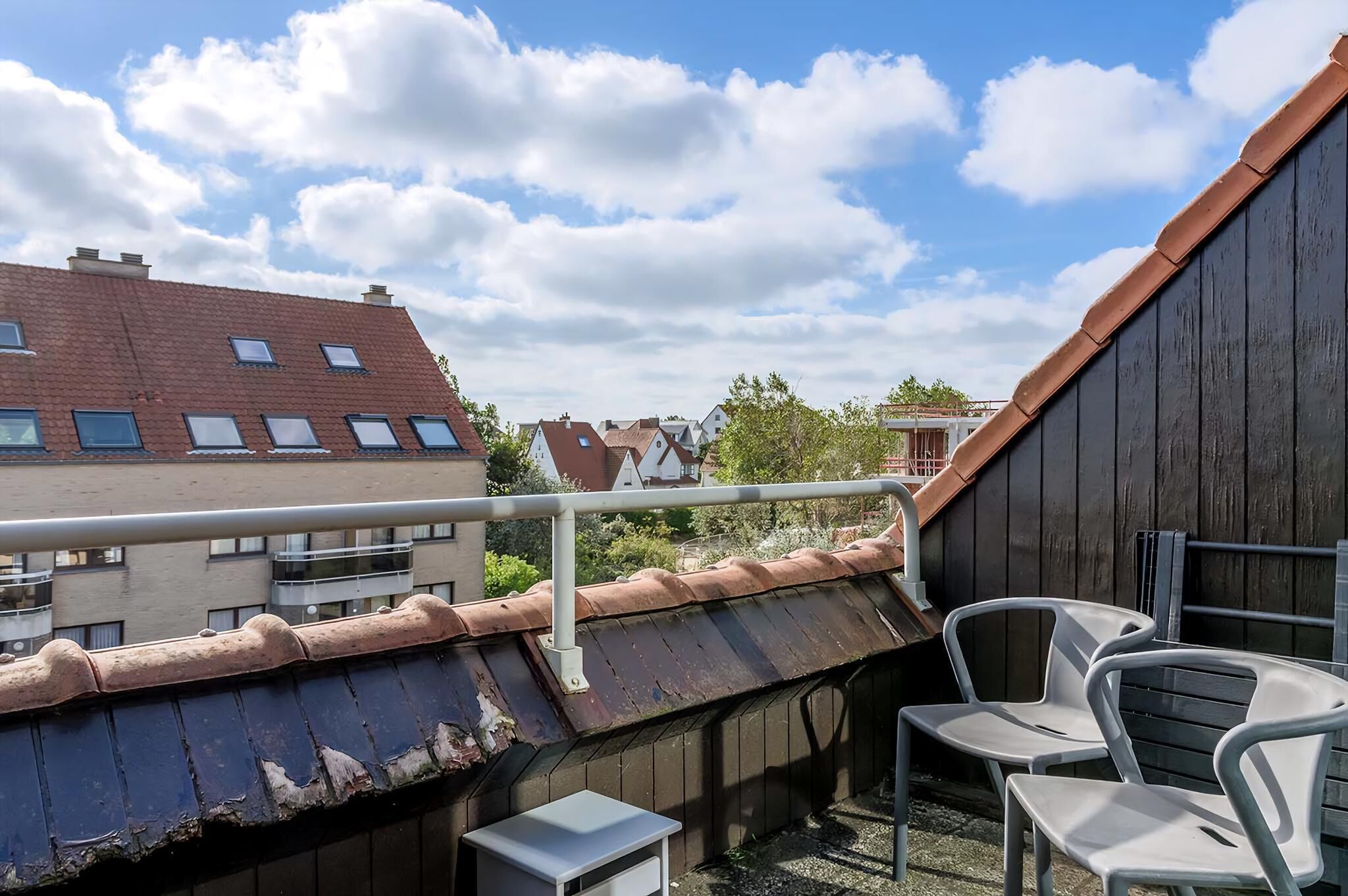 A rooftop terrace with a view of neighboring buildings, a railing, and two white plastic chairs.