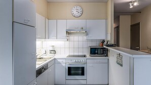 A compact kitchen with white cabinets, a built-in oven, a microwave, and a clock on the wall. - 2 bedroom accommodation in Oostduinkerke (Oostduinkerke)