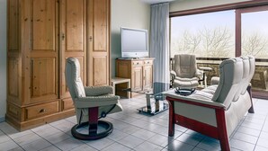 A room with a large wooden wardrobe, a television on a cabinet, a swivel chair, a sofa, and a balcony with a view of trees. - 2 bedroom accommodation in Oostduinkerke (Oostduinkerke)