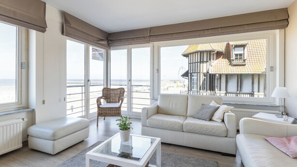 A spacious living room with a large window, a white sofa, a glass coffee table, and a wicker chair.