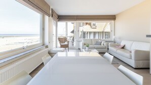 A spacious living room with a large window, a white sofa, a dining table, and a view of the sea.