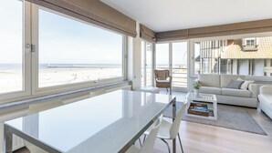 A modern living room with a glass dining table, white chairs, a sofa, and a view of the beach.