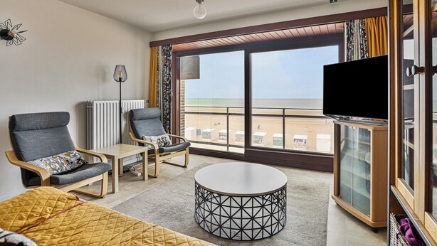 Een moderne hotelkamer met een groot raam, een ronde salontafel, twee fauteuils, een televisie en een kast.
