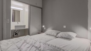 Makuuhuone, jossa on sänky, valkoiset vuodevaatteet, yöpöytä, peili ja seinään kiinnitetty valaisin.