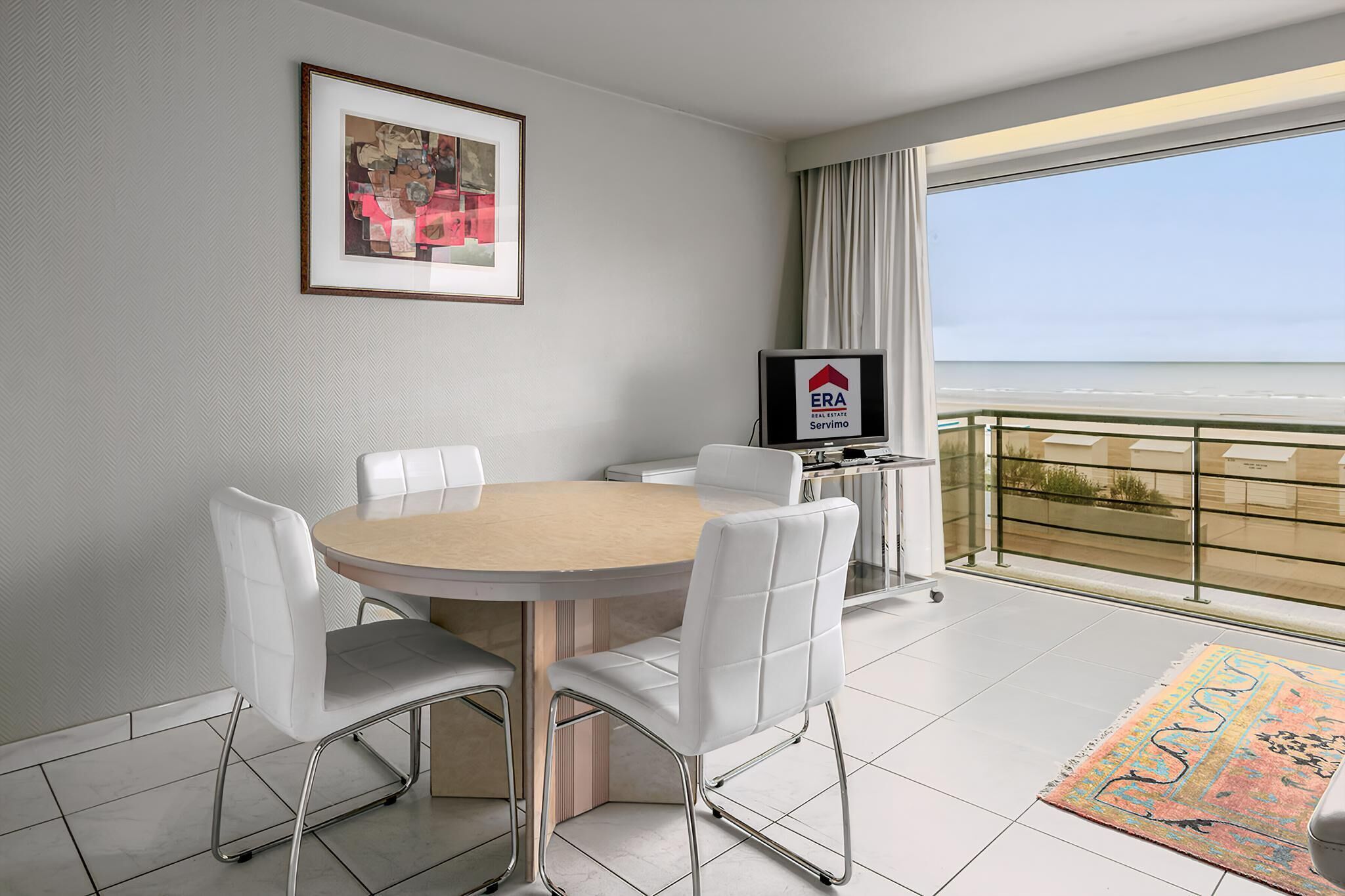Een hotelkamer met een ronde tafel, witte stoelen, een foto in een lijst aan de muur, een televisie en een balkon met uitzicht op het strand.