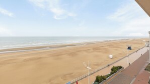 Une vue sur la plage depuis un balcon, avec un ciel dégagé, une plage de sable et une promenade bordée de lampadaires et d’une petite structure bleue.