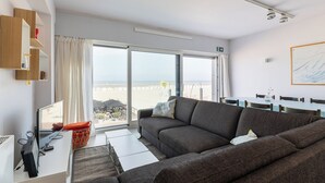 A modern living room with a large window, a sofa, a dining table, and a TV. - 3 bedroom accommodation in Oostduinkerke (Oostduinkerke)