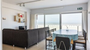 A room with a sofa, dining table, chairs, a TV, and a view of the beach. - 3 bedroom accommodation in Oostduinkerke (Oostduinkerke)