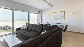 A modern living room with a large sofa, a dining table with chairs, and a view of the beach through large windows. - 3 bedroom accommodation in Oostduinkerke (Oostduinkerke)
