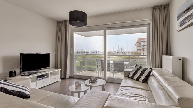 A modern living room with a flat-screen TV, a sofa, a coffee table, and a view of a balcony with outdoor furniture. - Villa Emilia 0206 + P104* (Nieuwpoort)