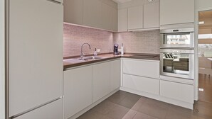 A modern kitchen with white cabinets, a brick backsplash, stainless steel appliances, and a large window. - Villa Emilia 0206 + P104* (Nieuwpoort)