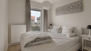 A bedroom with a large bed, a window with curtains, a radiator, a bedside table with a lamp, and a wall-mounted artwork. - Villa Emilia 0206 + P104* (Nieuwpoort)