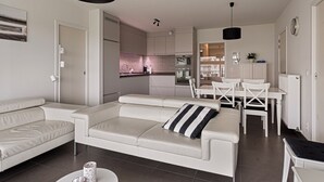 A modern living room with a white sofa, a dining table with chairs, and a kitchen in the background. - Villa Emilia 0206 + P104* (Nieuwpoort)
