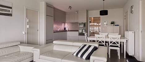 Een moderne woonkamer met een witte bank, een eettafel met stoelen en een keuken op de achtergrond.