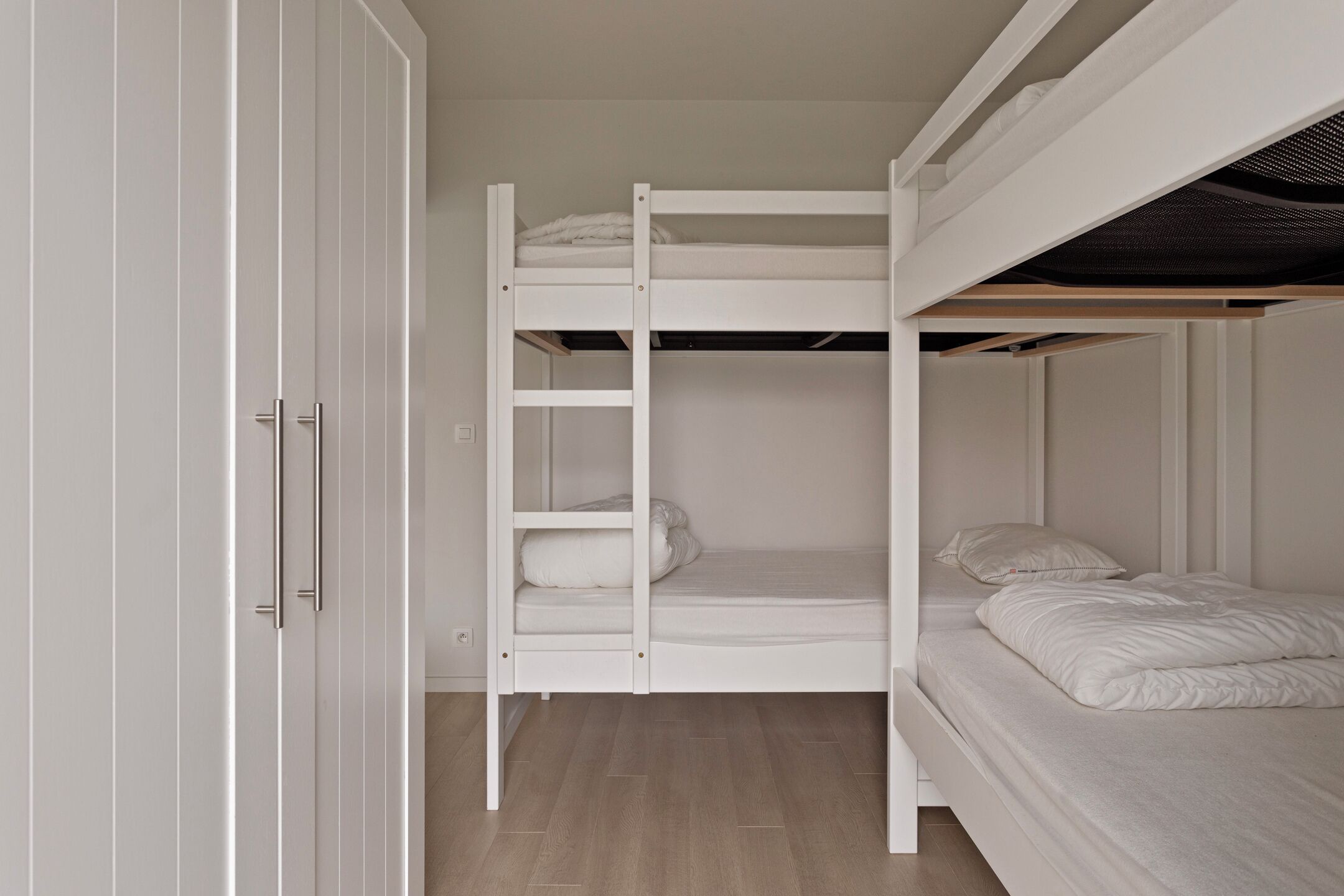Una cama nido con estructura y ropa de cama blancos, un armario con pomo y suelo de madera.