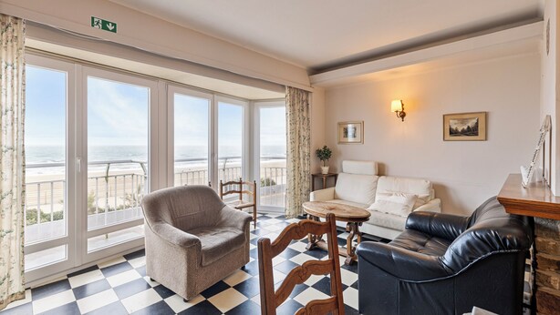 A room with a checkered floor, a sofa, a chair, a small table, and a view of the sea through large windows. - 2 bedroom accommodation in Koksijde (Koksijde)